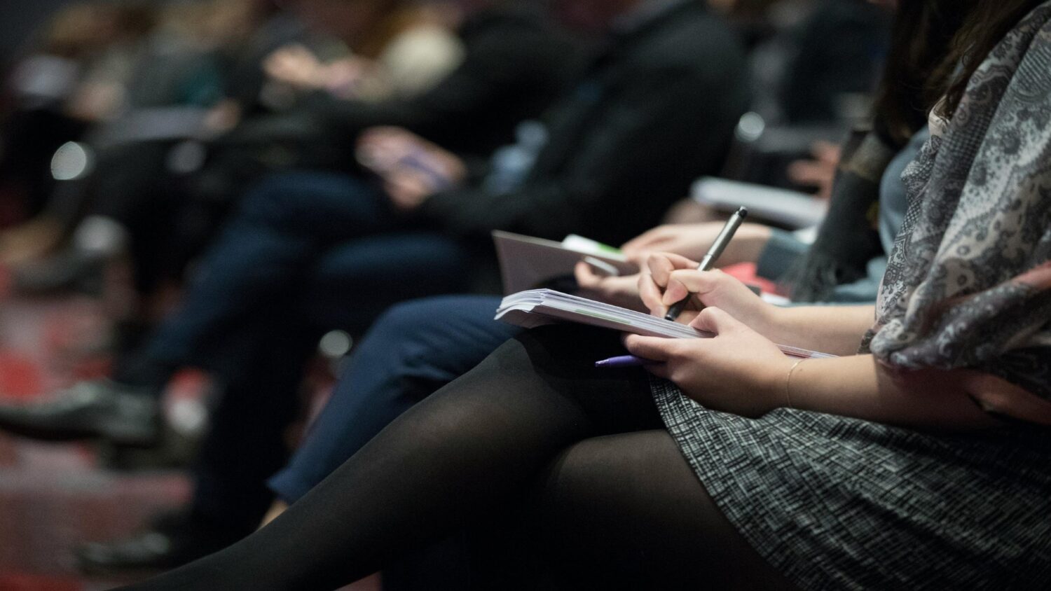 Decorative photo of a row of people seated in a room, taking notes on paper